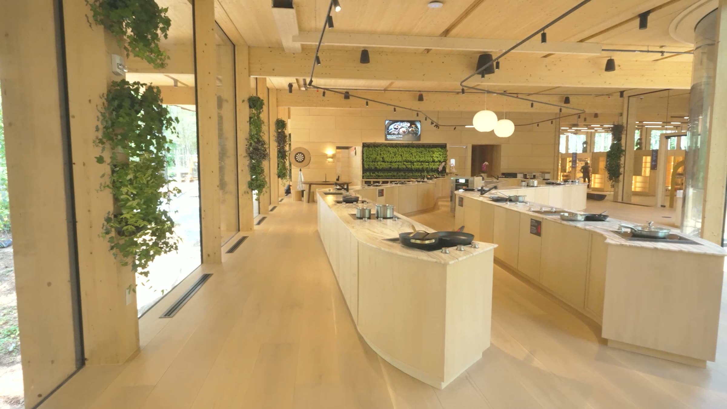 The Experiential Kitchen — the long curved cooking islands in pale timber, pans on the induction tops, the living wall and forest beyond