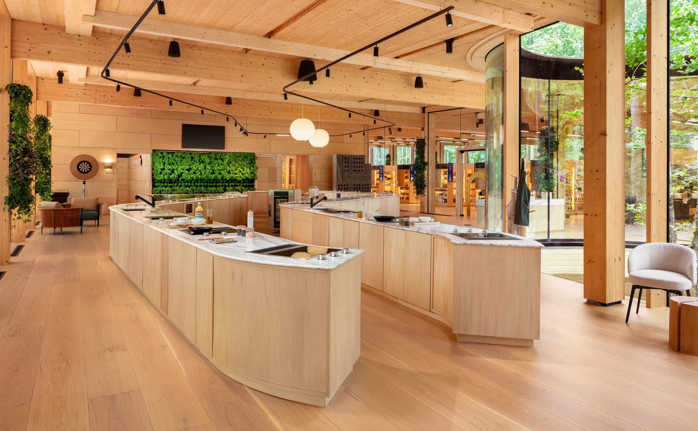 The Experiential Kitchen — two long curved cooking islands in pale timber, the living wall at the far end and the forest through the glass