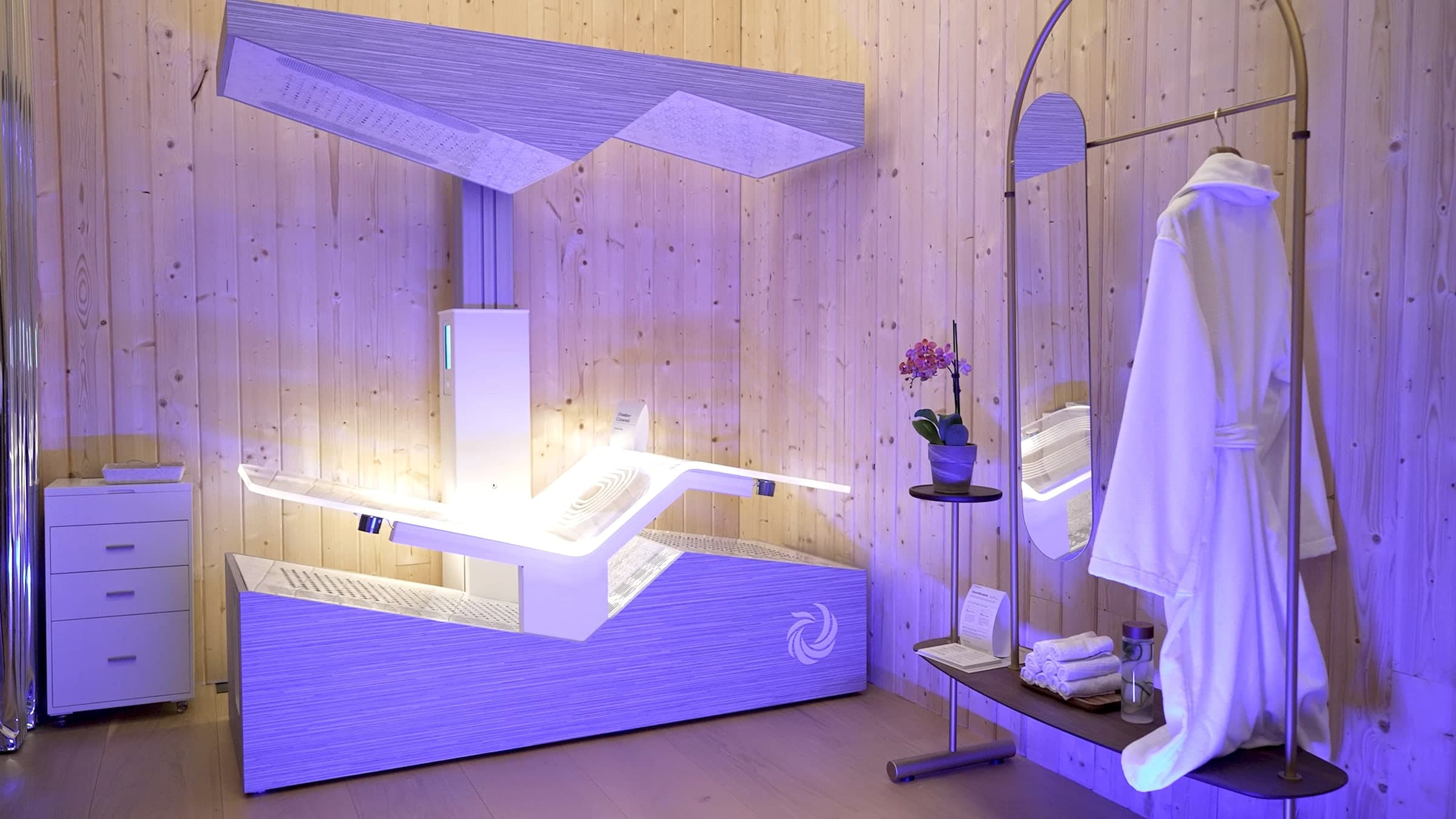 A Circadium therapy room — the sculpted white bed under its canopy in violet light, a robe hung beside the mirror